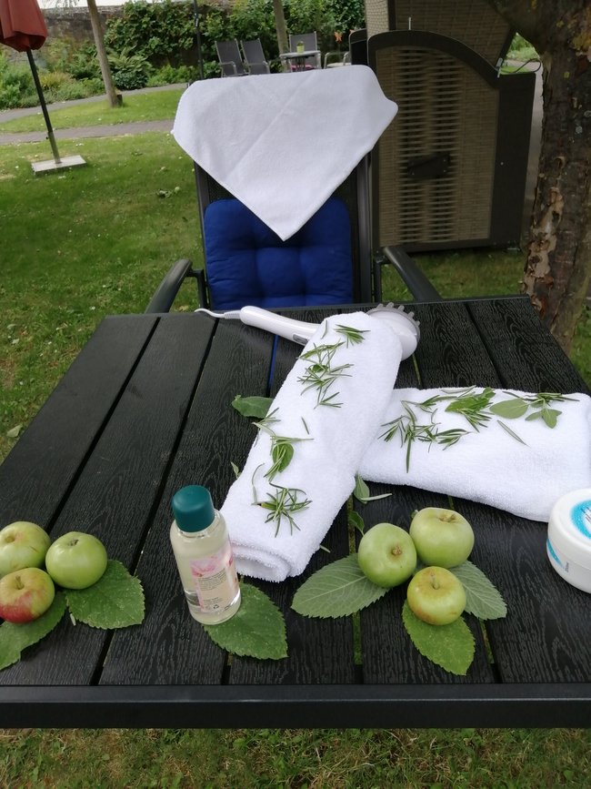 Wellness im Garten - der Gartentisch ist schön dekoriert mit Handtücher, Pflegecreme, Massagegerät und dekorativen Äpfeln.
