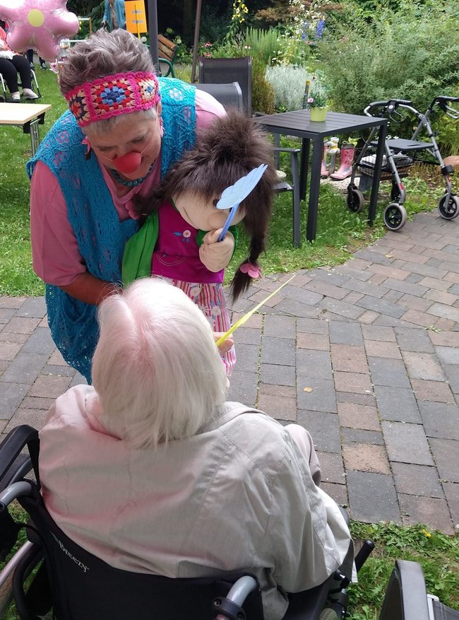 Clown mit Handpuppe bringt Seniorin zum Staunen.